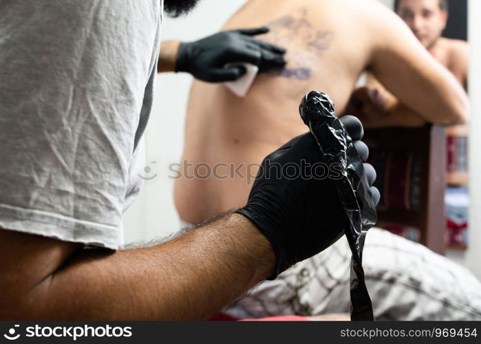 Professional master tattoo artist drawing tattoos picture on man's back using machine wearing black gloves at studio salon body art