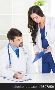 Professional medical doctor and his assistant working in the office, she is holding a clipboard and he is signing medical records. Medical doctor and his assistant