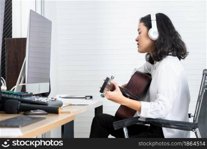 Professional musician young Asian woman vocalist Wearing Headphones plays guitar recording a song of the computer monitor of Software for recording and editing sounds in a professional studio