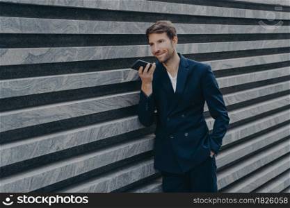 Professional prosperous adult man with stubble uses loudspeaker answers incoming call leaves voice audio message holds modern mobile phone near mouth dressed in formal suit poses near marble wall. Professional prosperous adult man with stubble uses loudspeaker answers incoming call
