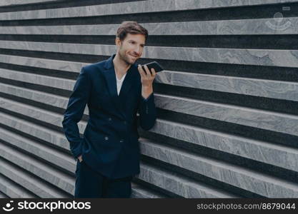 Professional prosperous adult man with stubble uses loudspeaker answers incoming call leaves voice audio message holds modern mobile phone near mouth dressed in formal suit poses near marble wall. Professional prosperous adult man with stubble uses loudspeaker answers incoming call