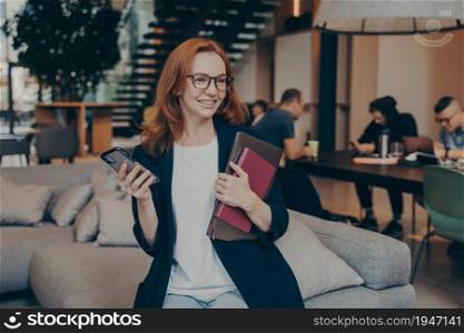 Professional smiling female worker with smartphone and laptop in hands waiting for colleague to arrive at meeting while sitting on couch in break room, dressed in casual clothes. Office life concept. Happy female worker waiting for her colleague to arrive while sitting on couch in office