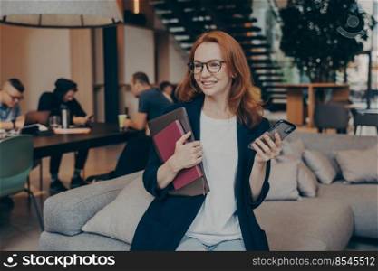Professional smiling female worker with smartphone and laptop in hands waiting for colleague to arrive at meeting while sitting on couch in break room, dressed in casual clothes. Office life concept. Happy female worker waiting for her colleague to arrive while sitting on couch in office