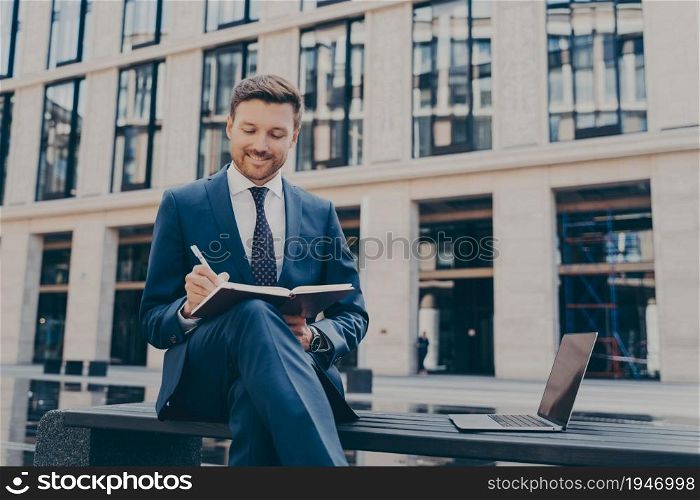 Professional smiling firm worker in formal suit writing down ideas with pen in note book while sitting on bench with his legs crossed, open laptop next to him, office buildings in blurred background. Professional office worker writing down ideas in notebook while working online outdoors