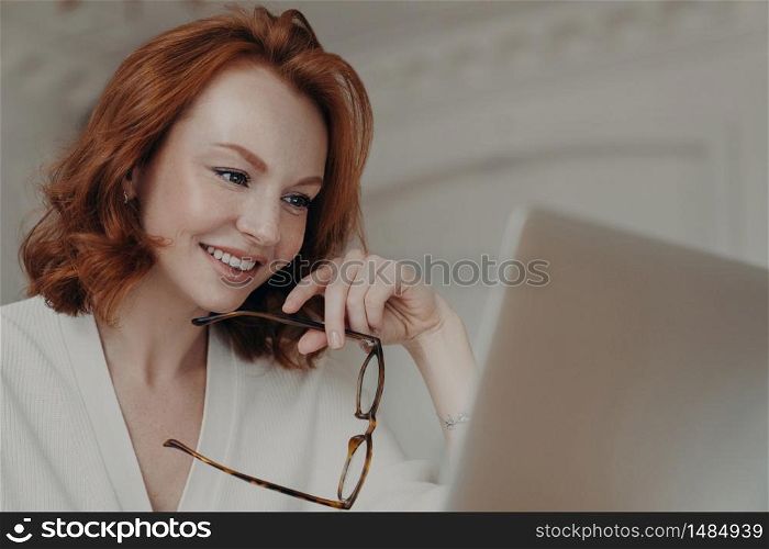 Professional smiling ginger female web designer uses application on laptop computer for creating project work, makes online research and browses internet, holds spectacles, has happy expression
