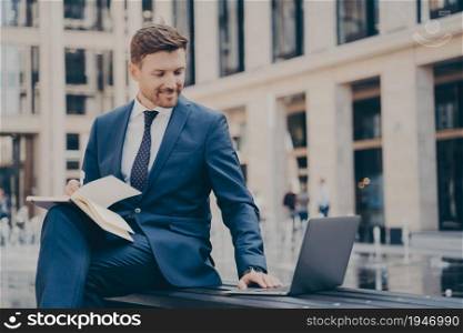 Professional young man company employee in blue formal suit doing research on laptop or watching business webinar, taking notes of important information in notebook, sitting on bench with legs crossed. Professional young company employee doing research on laptop or watching business webinar outdoors