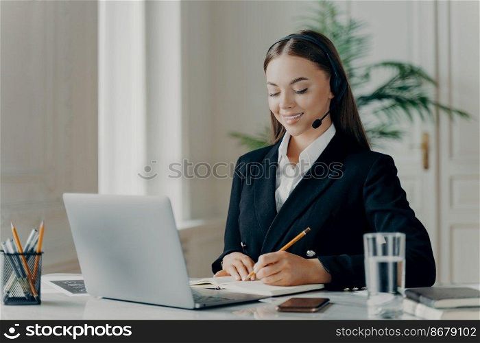 Proffesional female business consultant in formal suit and headset making notes and feeling sitisfied during web conference on laptop, manager of call center sitting in front of laptop at office. Smiling young businesswoman looking down and writing notes