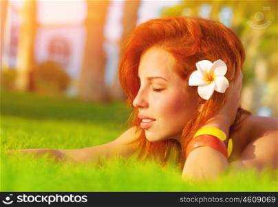 Profile of attractive woman with closed eyes dreaming outdoors, lying down on fresh green grass glade, frangipani flower in red hair, enjoying day spa, luxury summer resort