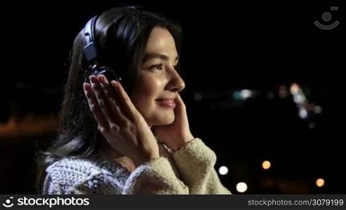 Profile of beautiful young brunette woman with headphones enjoying favorite song while relaxing at night city. Smiling charming girl holding both hands on earcups while listening music with headphones at night over colorful bokeh of streetlights.