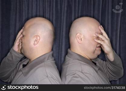 Profile of Caucasian bald identical twin men standing back to back and grimicing with hands to head.