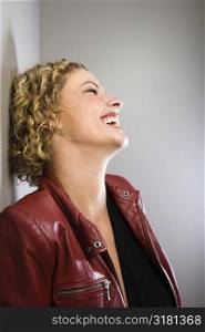 Profile of Caucasian young adult woman in red jacket leaning back against wall laughing.