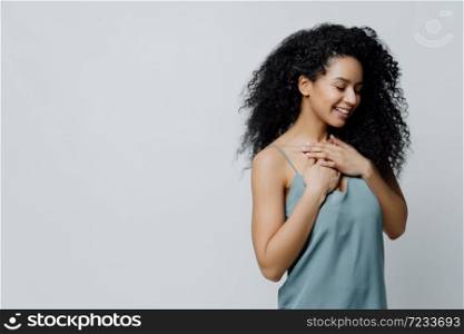 Profile of dreamy romantic ethnic woman with Afro haircut, dressed in nightwear, keeps hands on chest, recalls very pleasant moment in life smiles tenderly with closes eyes expresses truthful feelings