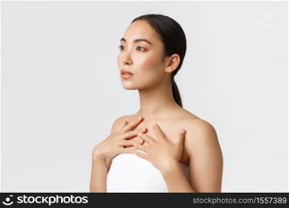 Profile of feminine sensual young asian woman in white towel looking upper left corner and touching her body, promo of cosmetics, skincare products or face creams, white background.. Profile of feminine sensual young asian woman in white towel looking upper left corner and touching her body, promo of cosmetics, skincare products or face creams, white background