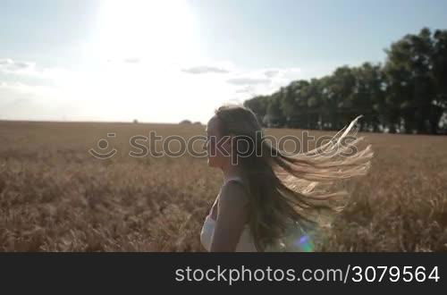 Profile of happy young blond female with flying long hair running through golden wheat field in summer against blue sky background. Positive free beautiful woman enjoying nature, running, jumping, spinning around in cereal field on countryside.