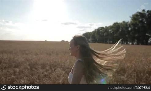 Profile of happy young blond female with flying long hair running through golden wheat field in summer against blue sky background. Positive free beautiful woman enjoying nature, running, jumping, spinning around in cereal field on countryside.