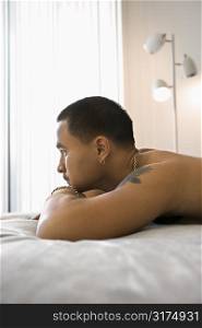Profile of shirtless Asian young adult man lying on bed resting head on arms looking out window.
