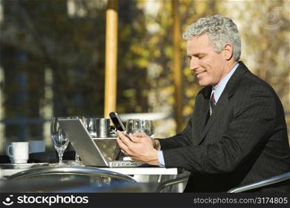 Profile of smiling prime adult Caucasian man in suit sitting at patio table outside with laptop and dialing cellphone.