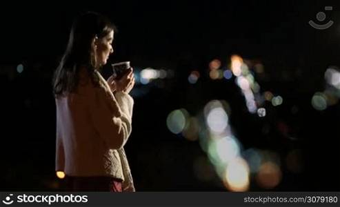 Profile of young beautiful brunette female enjoying night city lights while standing on observation deck and drinking coffee. Attractive millennial woman with coffee to go relaxing at night in park against colorful bokeh background
