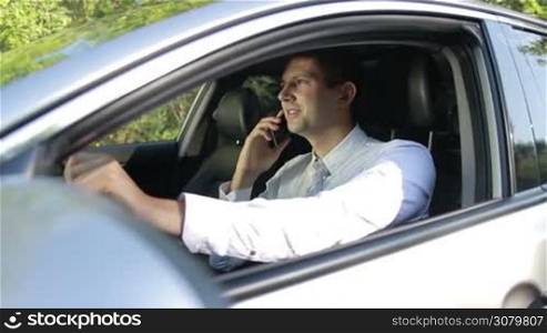 Profile of young emotional businessman in white shirt sitting in car, arguing with foreign partner using smartphone and gesturing. Serious entrepreneur talking on mobile phone while driving car. Side view. Slow motion. Steadicam stabilized shot.