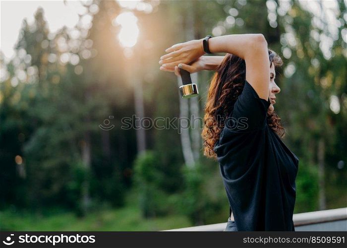Profile shot of active motivated young woman dressed in black t shirt, raises dumbbell over head, poses against trees with sunset, has workout outdoor, wants to have muscular arms. Sport concept