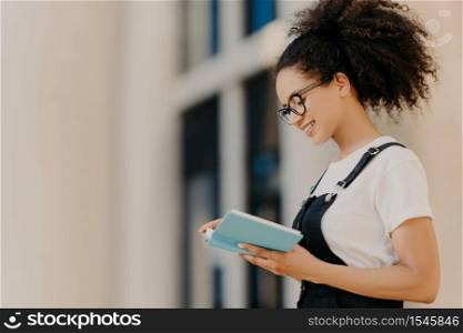 Profile shot of curly girl focused in organizer, learns her plans for next week, has positive expression, wears optical glasses, white t shirt and overalls. African American woman with diary outdoor