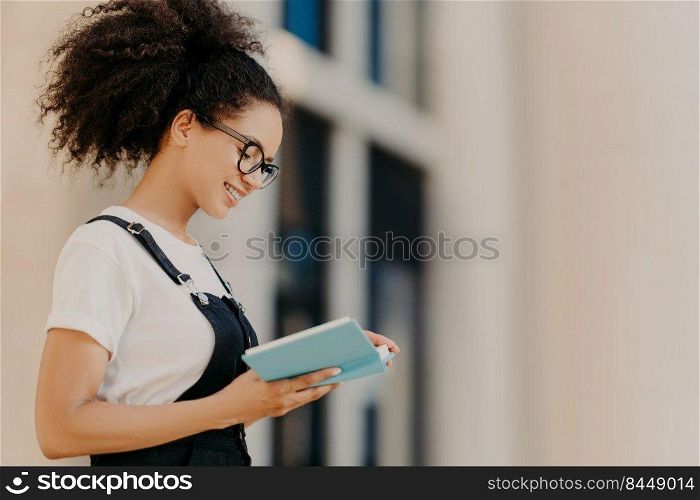 Profile shot of curly girl focused in organizer, learns her plans for next week, has positive expression, wears optical glasses, white t shirt and overalls. African American woman with diary outdoor