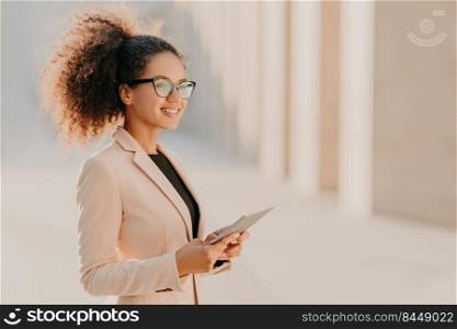 Profile shot of glad dark skinned woman with Afro hair, dressed elegantly holds tablet computer in hands strolls at street wears optical glasses connected to high speed internet. Lifestyle, technology