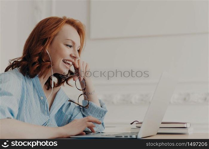 Profile shot of happy redhead woman laughs positively, has online communication with friend, takes break after work, wears blue shirt, poses in coworking space. People, freelance work, distance job