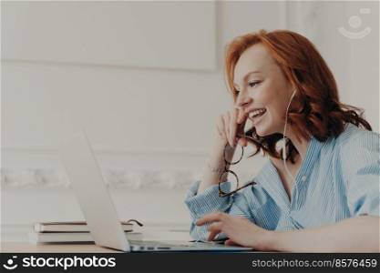 Profile shot of happy redhead woman laughs positively, has online communication with friend, takes break after work, wears blue shirt, poses in coworking space. People, freelance work, distance job