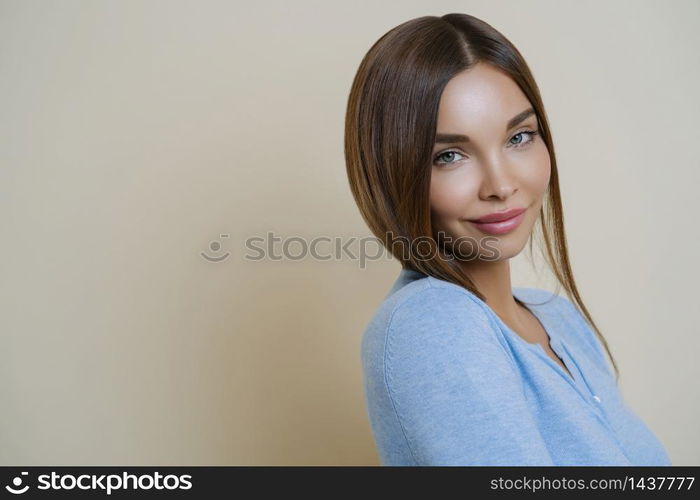 Profile shot of lovely dark haired woman with European appearance, has healthy well cared skin, minimal makeup, wears blue jumper, stands against beige background, copy space for your promotion