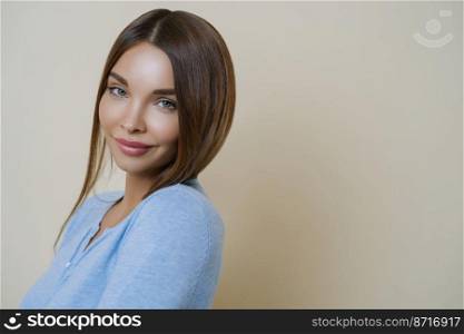 Profile shot of lovely dark haired woman with European appearance, has healthy well cared skin, minimal makeup, wears blue jumper, stands against beige background, copy space for your promotion