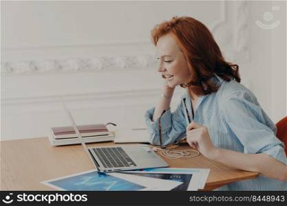 Profile shot of pleased redhead woman entrepreneur enjoys video conference with colleague, poses at desktop with paper documents, pleased to work from home, glad to see friend. Vitrual reality