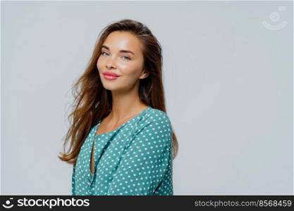 Profile shot of pretty pleased woman wears makeup, rosy lipstick, dressed in polka dot blouse, has healthy skin, poses against white background with blank space for your advert or promotion.