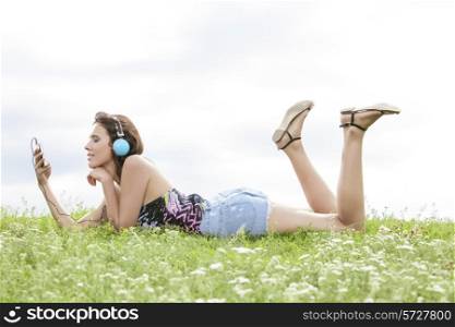 Profile shot of woman listening to music through cell phone using headphones while lying on grass against sky