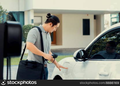 Progressive asian man install cable plug to his electric car with home charging station in the backyard. Concept use of electric vehicles in a progressive lifestyle contributes to clean environment.. Progressive asian man recharge his EV car at home charging station.