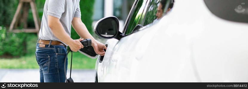 Progressive asian man install cable plug to his electric car with home charging station in the backyard. Concept use of electric vehicles in a progressive lifestyle contributes to clean environment.. Progressive asian man recharge his EV car at home charging station.