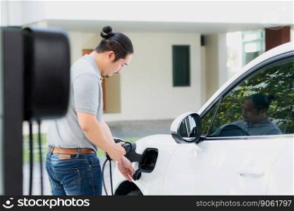 Progressive asian man install cable plug to his electric car with home charging station in the backyard. Concept use of electric vehicles in a progressive lifestyle contributes to clean environment.. Progressive asian man recharge his EV car at home charging station.