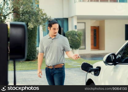 Progressive asian man install cable plug to his electric car with home charging station in the backyard. Concept use of electric vehicles in a progressive lifestyle contributes to clean environment.. Progressive asian man recharge his EV car at home charging station.