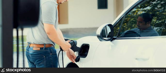 Progressive asian man install cable plug to his electric car with home charging station in the backyard. Concept use of electric vehicles in a progressive lifestyle contributes to clean environment.. Progressive asian man recharge his EV car at home charging station.