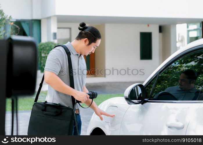 Progressive asian man install cable plug to his electric car with home charging station in the backyard. Concept use of electric vehicles in a progressive lifestyle contributes to clean environment.. Progressive asian man recharge his EV car at home charging station.