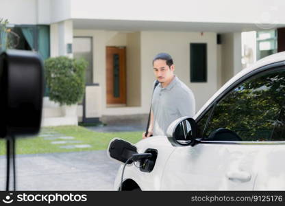 Progressive asian man install cable plug to his electric car with home charging station in the backyard. Concept use of electric vehicles in a progressive lifestyle contributes to clean environment.. Progressive asian man recharge his EV car at home charging station.