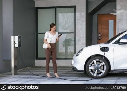 Progressive asian woman holding smartphone with electric car at home charging station. Concept of the use of electric vehicles in a progressive lifestyle contributes to a clean and healthy environment. Progressive woman holding phone at EV home charging station in garage.