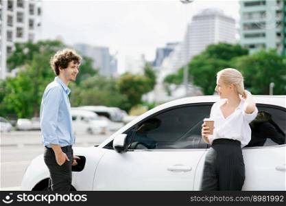 Progressive businessman and businesswoman with coffee, standing at electric car connected to charging station before driving around city center. Eco friendly rechargeable car powered by clean energy.. Progressive businessman and businesswoman with coffee at public charging station