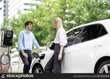 Progressive businessman and businesswoman with electric car parking and connected to public charging station before driving around city center. Eco friendly rechargeable car powered by clean energy.. Progressive businessman and businesswoman at charging point and EV car