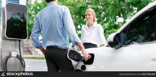 Progressive businessman and businesswoman with electric car parking and connected to public charging station before driving around city center. Eco friendly rechargeable car powered by clean energy.. Progressive businessman and businesswoman at charging point and EV car