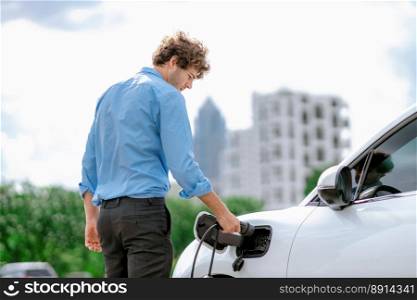 Progressive businessman insert charger plug from charging station to his electric vehicle with apartment condo building in background. Eco friendly rechargeable car powered by sustainable energy.. Progressive businessman plugs charger plug from charging station to EV.