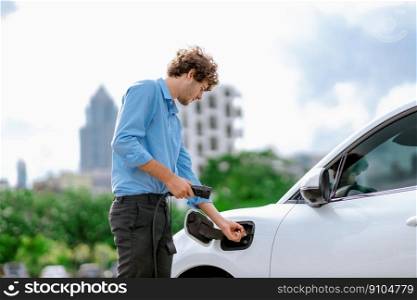 Progressive businessman insert charger plug from charging station to his electric vehicle with apartment condo building in background. Eco friendly rechargeable car powered by sustainable energy.. Progressive businessman plugs charger plug from charging station to EV.