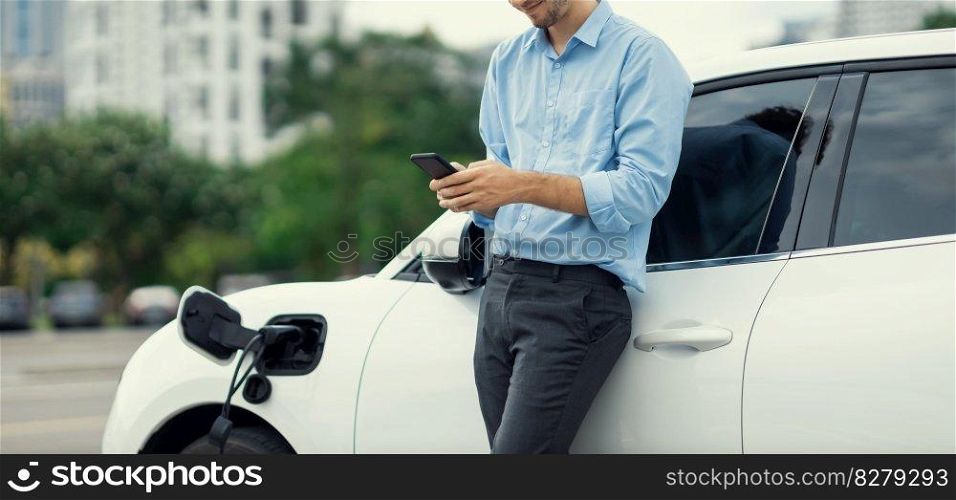 Progressive businessman talking on the phone, leaning on electric car recharging with public EV charging station, apartment condo residential building on the background as green city lifestyle.. Progressive businessman talking on the phone with recharging electric vehicle.