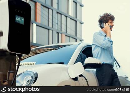 Progressive businessman talking on the phone, leaning on electric car recharging with public EV charging station, apartment condo residential building on the background as green city lifestyle.. Progressive businessman talking on the phone with recharging electric vehicle.