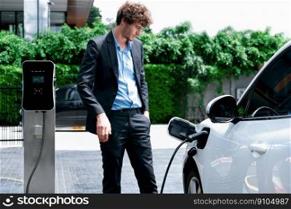 Progressive businessman wearing black suit with electric car recharging at public parking car charging station at modern city residential area. Eco friendly rechargeable car powered by clean energy.. Progressive businessman with EV car at public parking car charging station.